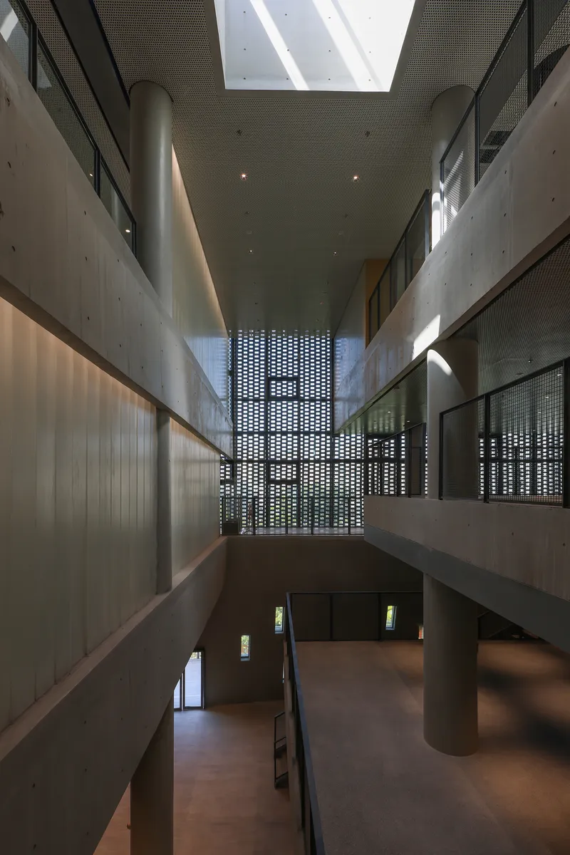Double-height atrium with board-formed concrete walls, perforated screen and diagonal skylight above cylindrical columns