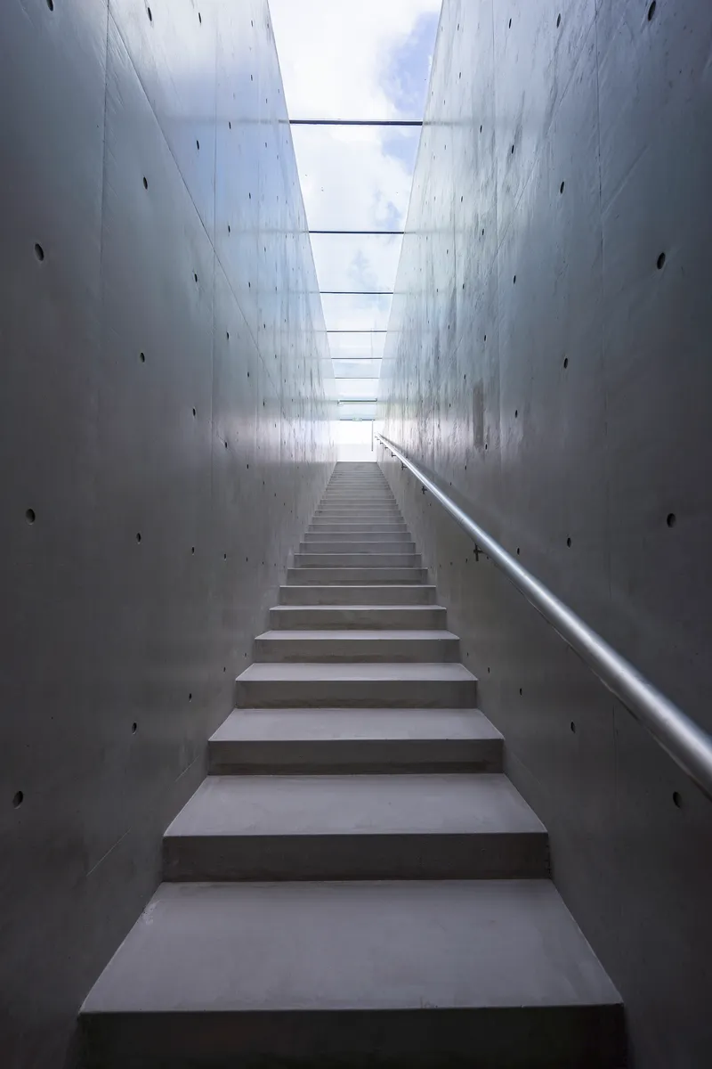 Concrete staircase ascending between tall cast-in-place walls beneath a linear skylight admitting daylight