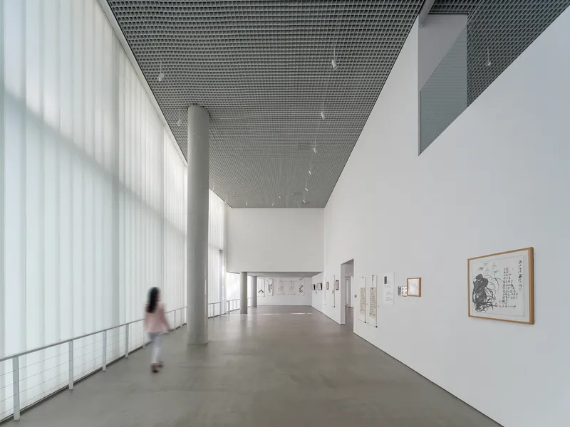 Gallery with perforated metal ceiling, translucent curtain wall and a figure walking past framed artworks