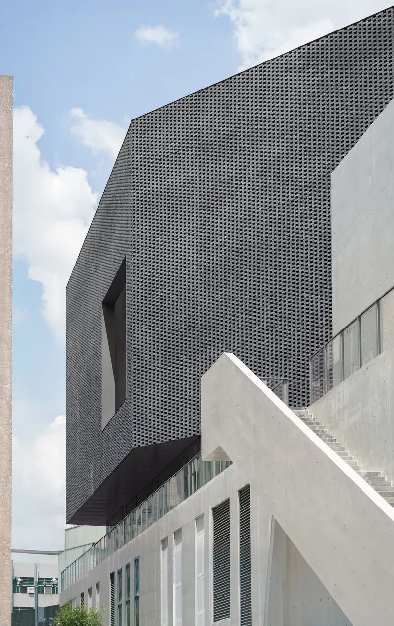 Perforated dark metal facade volume rising above white concrete buildings under scattered clouds
