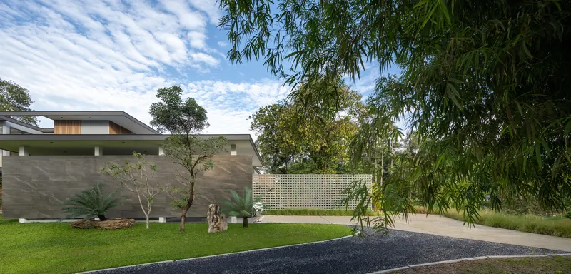 Gravel driveway leading to the stone-clad entrance wall and perforated screen beyond a lawn with trees