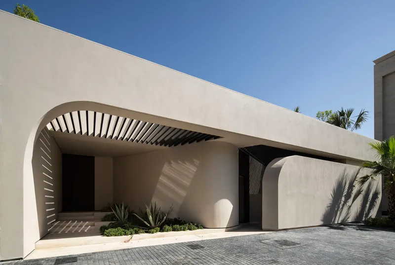Arched entry with radial timber slat canopy casting shadows on pale stucco wall and planted bed