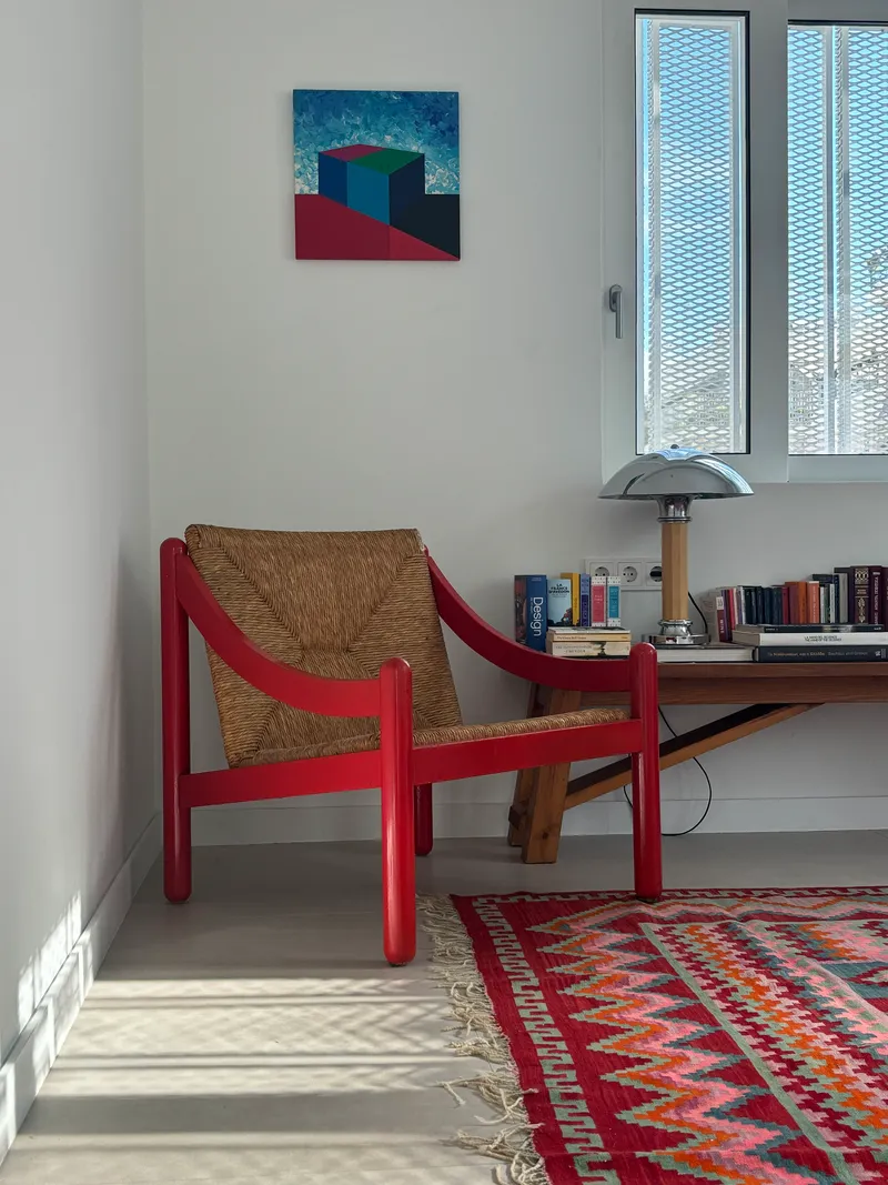 Living space corner with woven rush chair, low bookshelf, and narrow window with perforated shutters