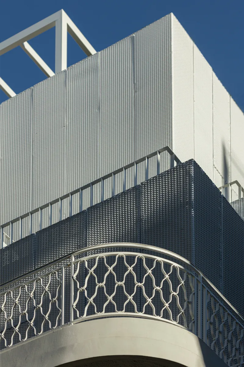 Curved terrace balcony with geometric metal railing below perforated white and dark cladding panels under clear sky
