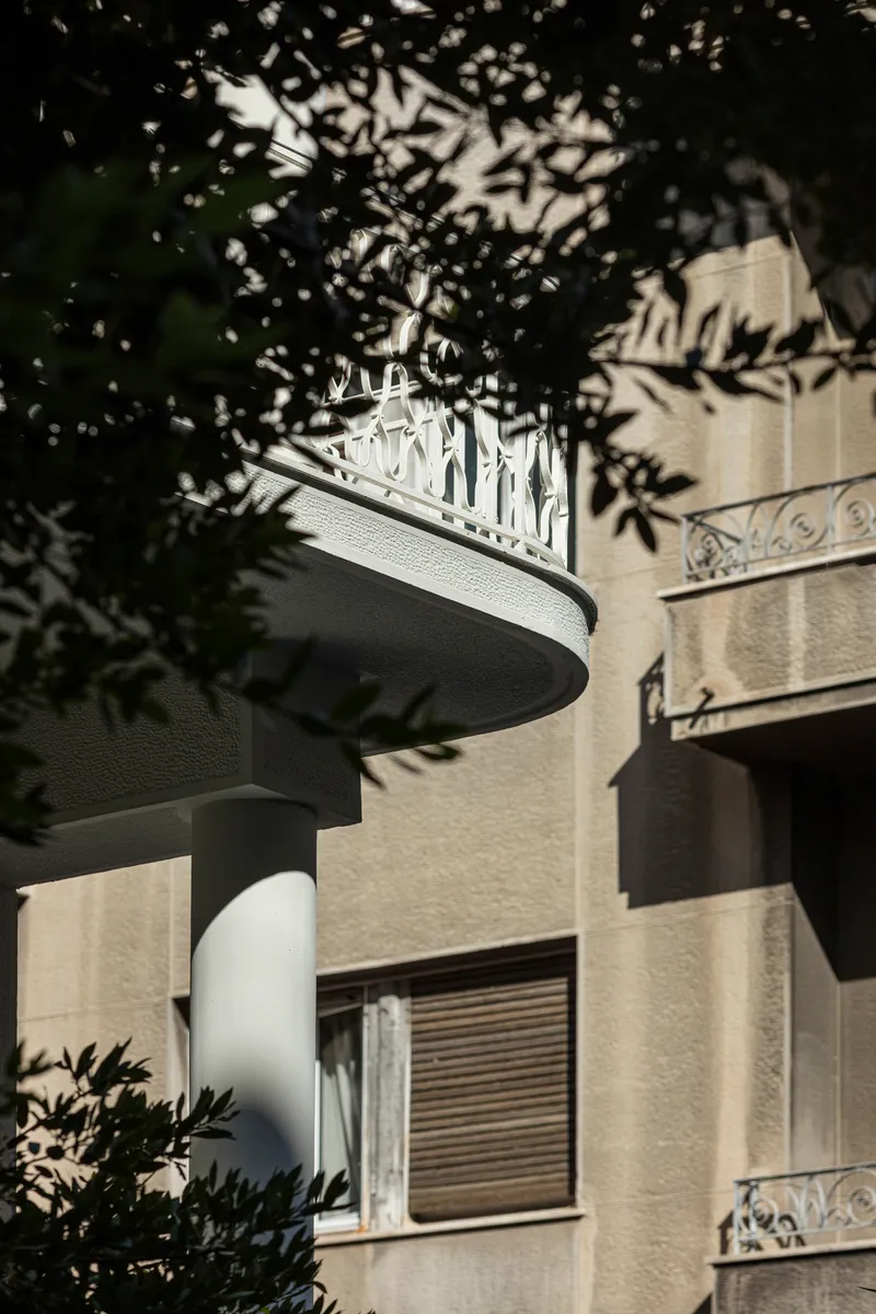 Curved balcony with decorative metal railing visible through dappled tree shadow cast on adjacent beige facade