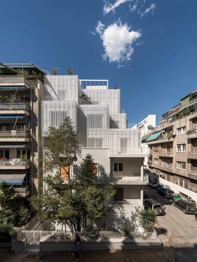 Street view of the white textured facade rising between planted trees under a clear blue sky