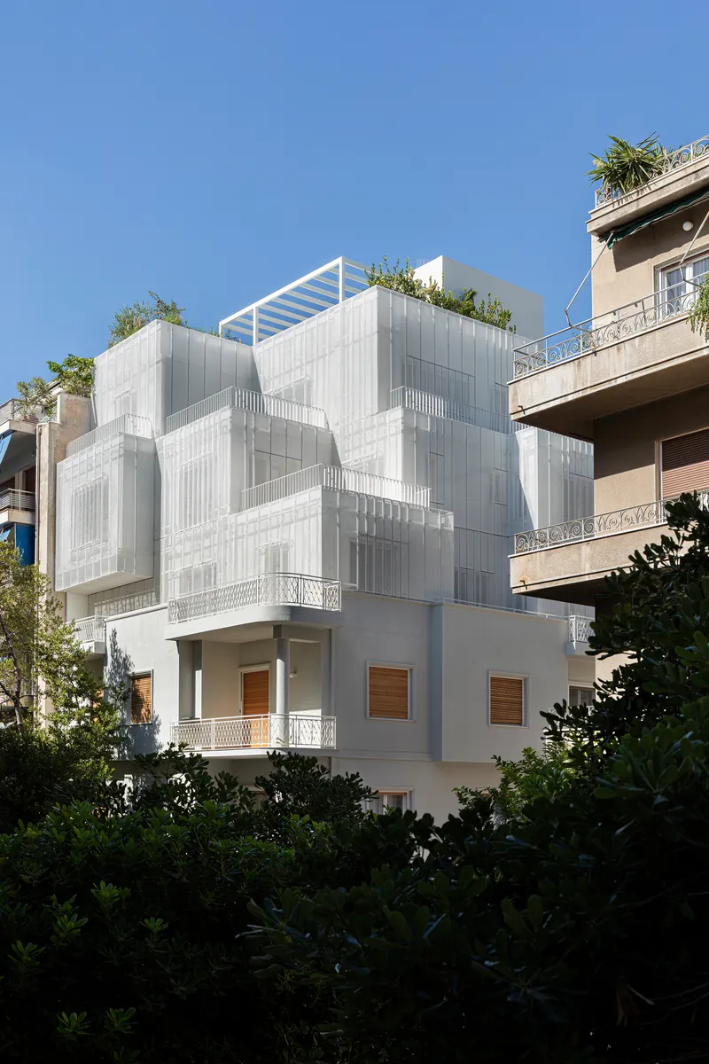 Street view of the layered white metal screen facade with rooftop garden framed by neighboring buildings