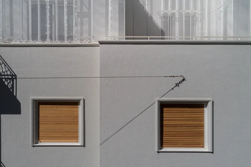 Two square windows with timber roller shutters set into pale rendered wall with diagonal shadow line