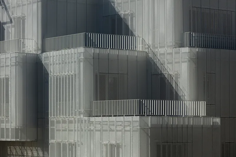 Offset balconies with slatted railings recessed into perforated metal screen facade with shadow patterns across surface