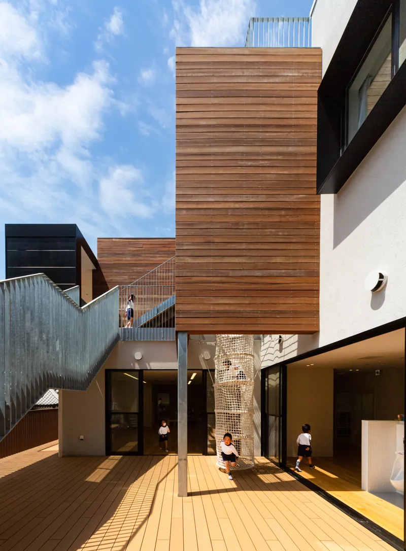 Timber-clad tower rising above a courtyard with decking and children playing in afternoon sunlight
