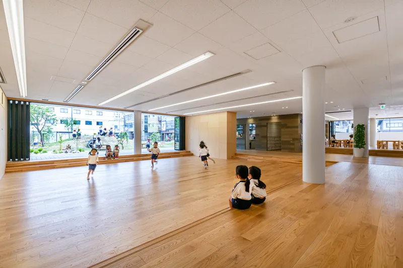 Open timber-floored hall with white cylindrical column and children playing near full-height glazing