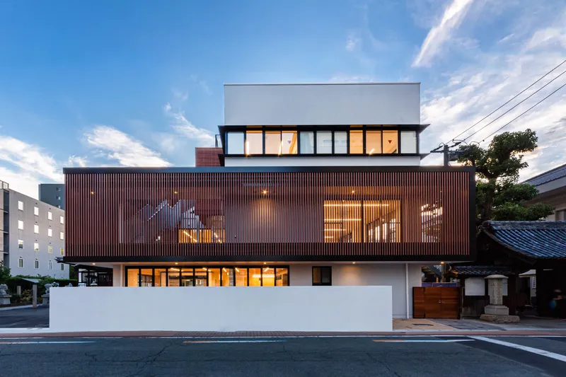 Street facade with vertical timber screen at middle level illuminated at dusk under cloudy sky