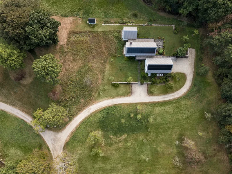 Aerial view of three gabled volumes with metal roofs arranged along a curving gravel path