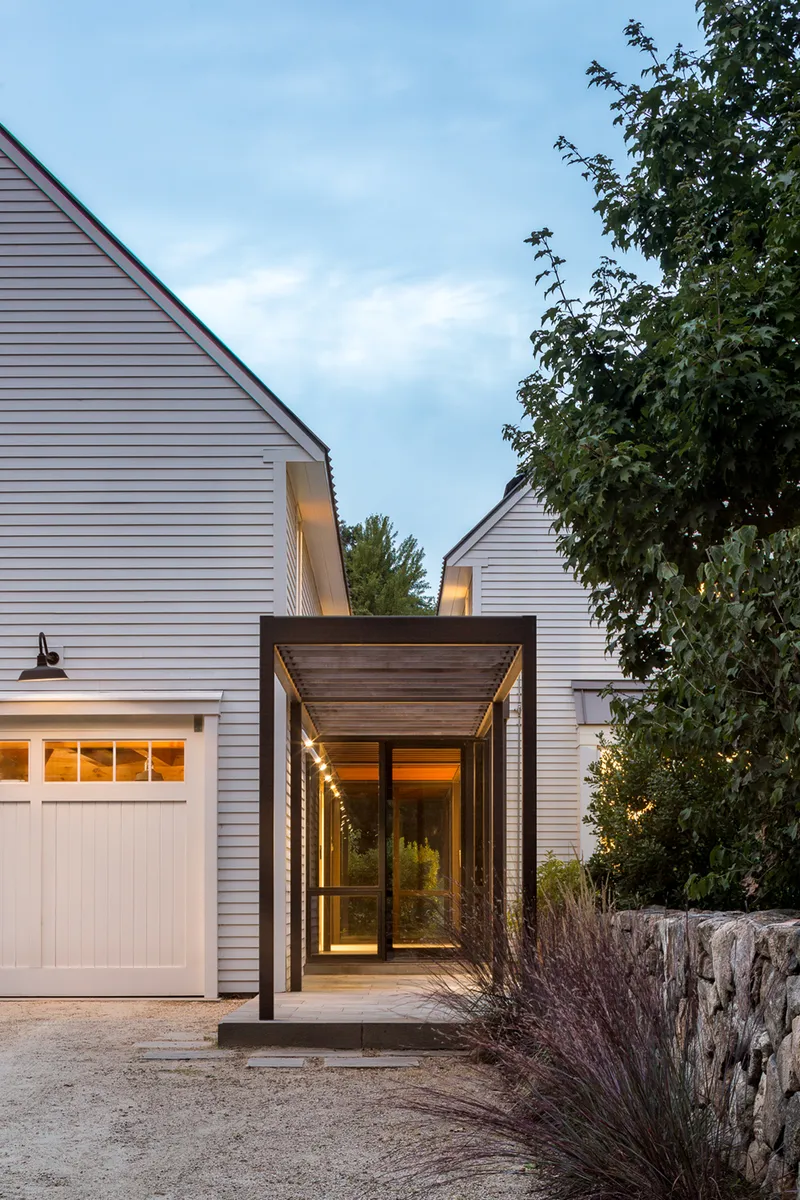 Timber-framed entry canopy connecting white horizontal siding volumes flanked by ornamental grasses at twilight