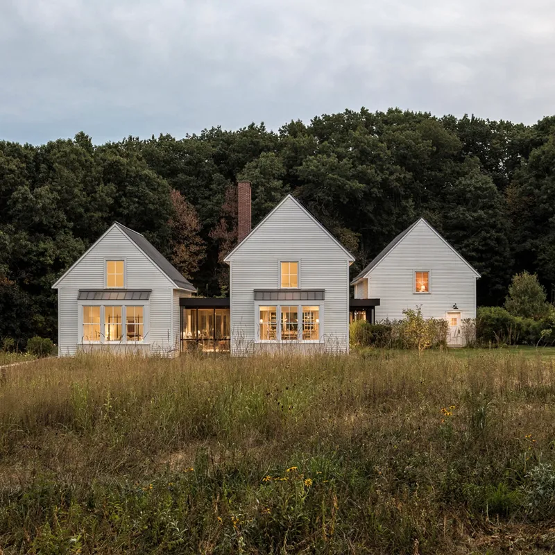 Three white-clad gabled volumes with lit windows across a meadow at dusk