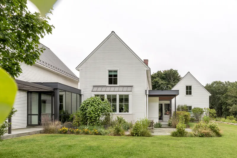 White horizontal siding and standing seam metal roofs across three gabled volumes framing native grass plantings