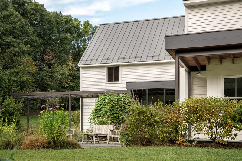 Standing seam metal roof over white clapboard siding with a timber pergola and garden beds