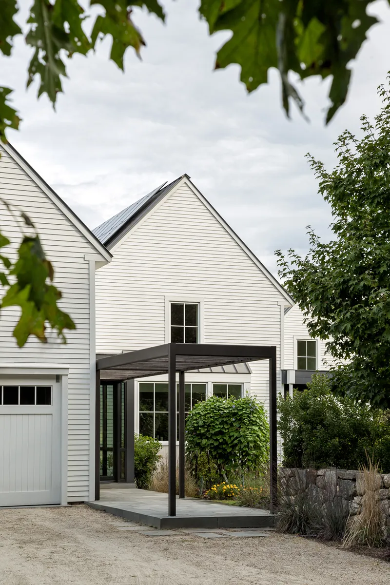Steel and timber canopy framing the entrance beneath white gabled volumes seen through tree branches