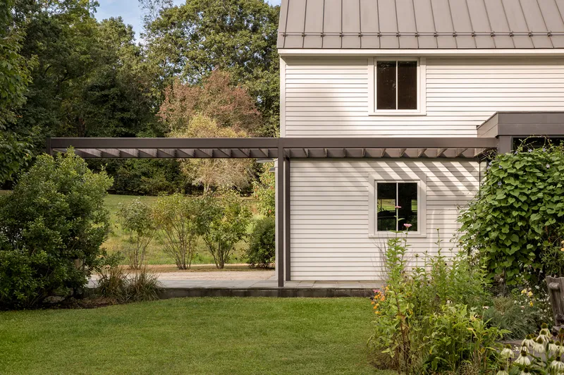 Timber pergola with exposed rafters casting shadow stripes across white clapboard facade and planted beds
