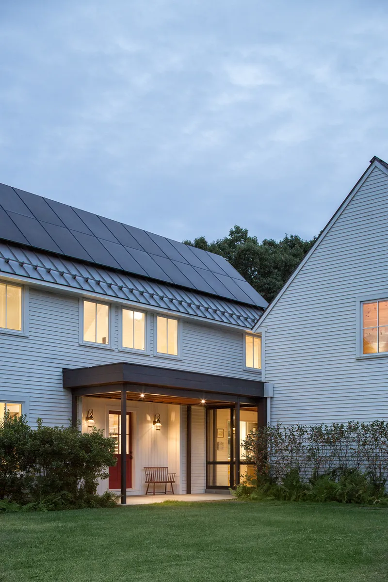 Covered porch with timber posts beneath white siding and solar panels on the gabled roof at dusk