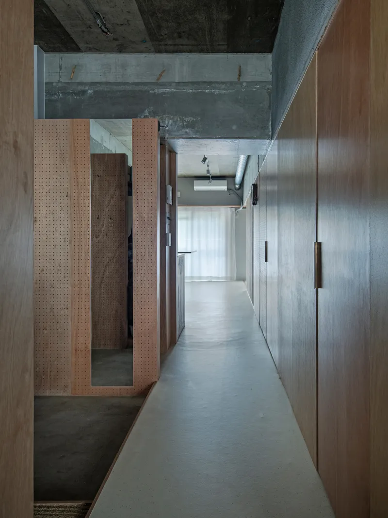 Narrow corridor with perforated timber panel and concrete walls leading toward a backlit curtained opening