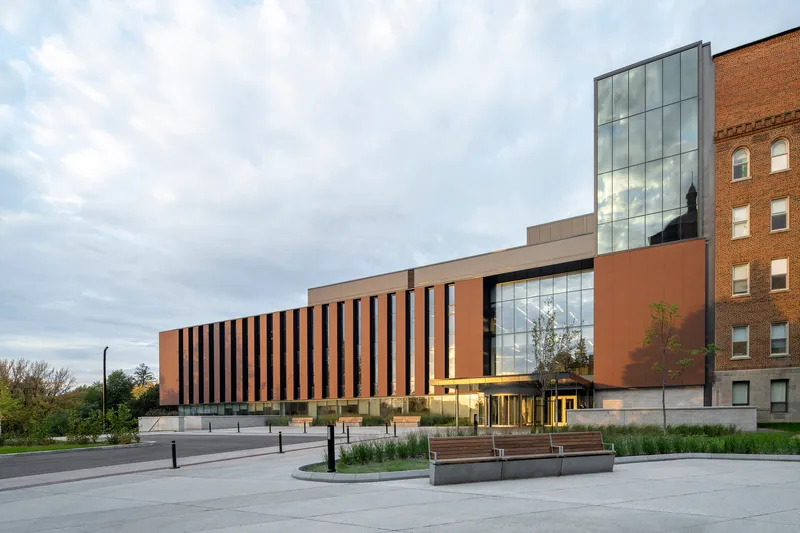 Street view of the copper-paneled facade with glazed entrance and concrete plaza benches under cloudy skies