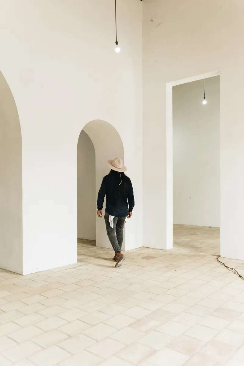 Person in hat walking through arched doorway connecting pale tiled rooms with pendant light bulbs