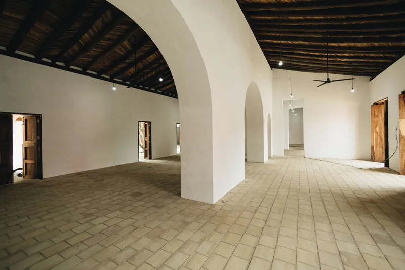 Interior gallery space with arched openings in white walls and woven bamboo ceiling above clay tile floor