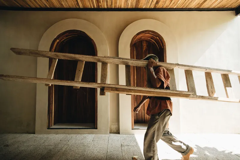 Person carrying a timber ladder past arched openings and plastered walls