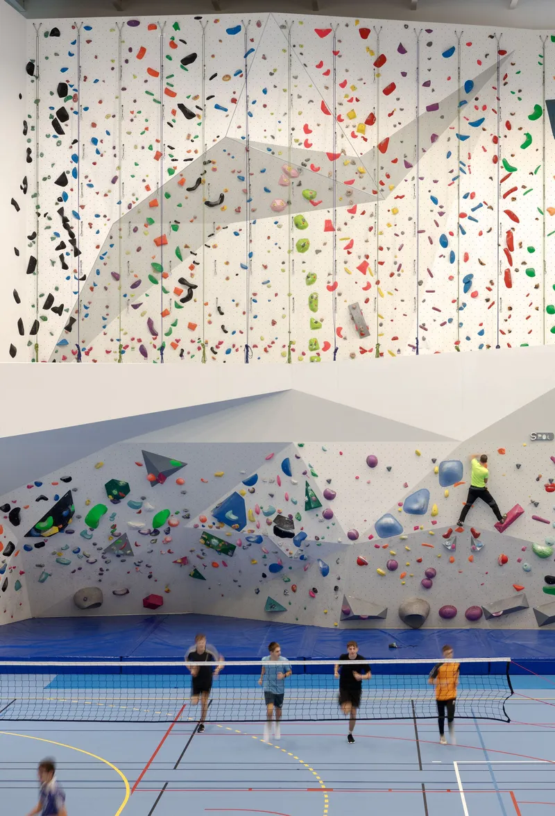 Four people standing at badminton net below two-level climbing walls with multicolored holds