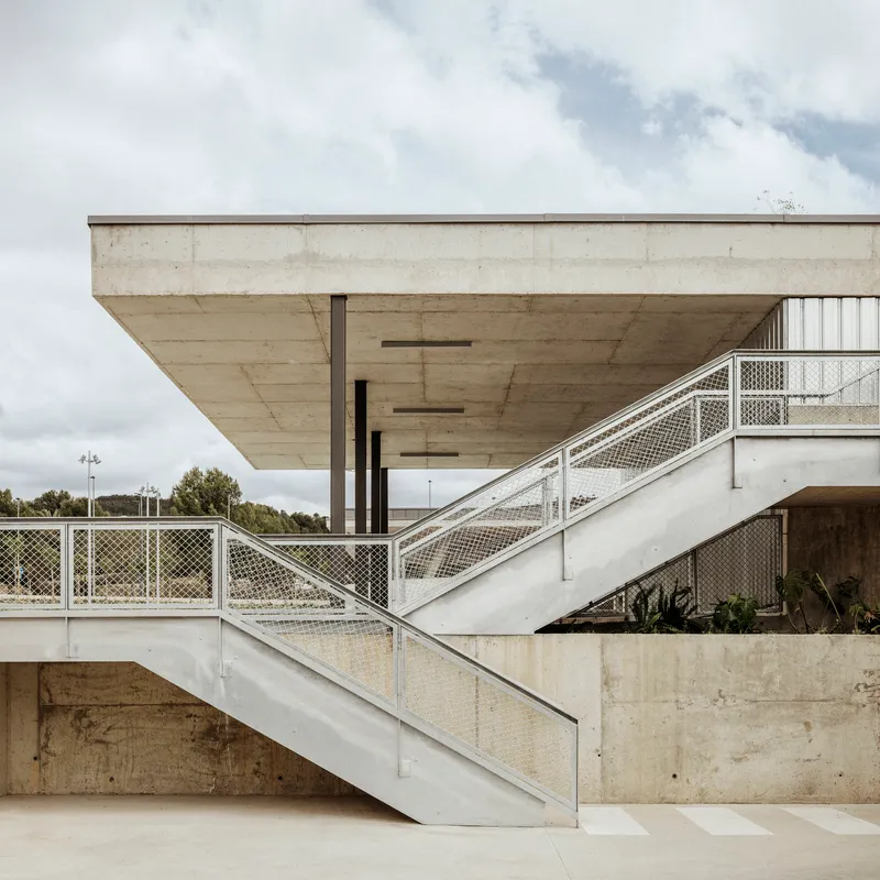 Exterior concrete stair with metal mesh railings ascending to a cantilevered soffit under cloudy sky