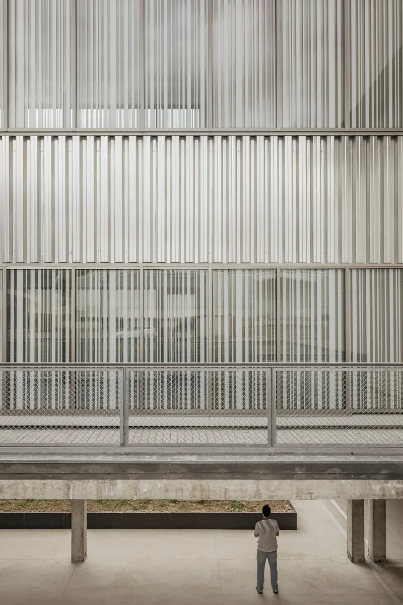 Vertical metal screen facade with mesh balustrade viewed from concrete-columned ground-level courtyard with visitor