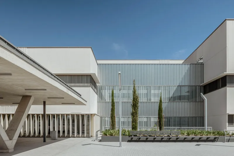 Courtyard with corrugated metal facade, vertical slat screening, planted beds with cypress trees, and concrete benches