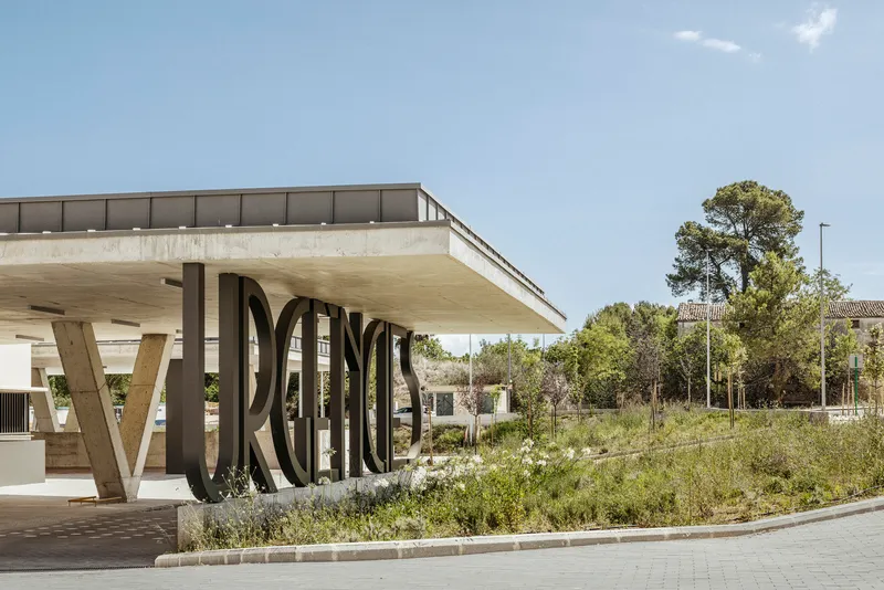 Cantilevered concrete canopy on angled supports with metal lettering and wildflower meadow under blue sky