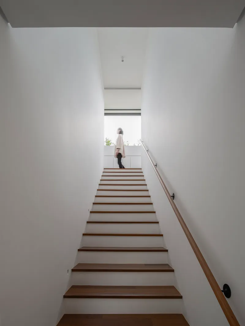 Timber staircase rising between white walls with a figure at the top landing