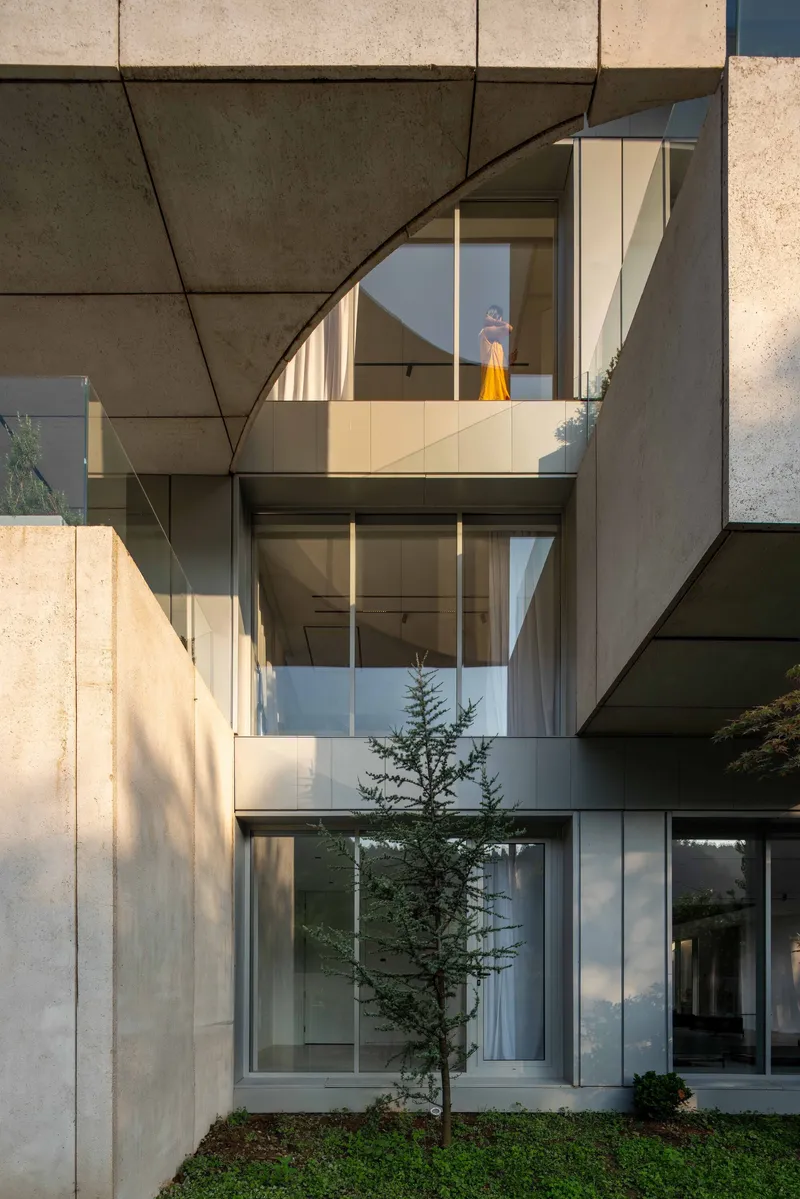 Courtyard view with stacked glass-enclosed levels, a young evergreen tree, and a figure in afternoon light