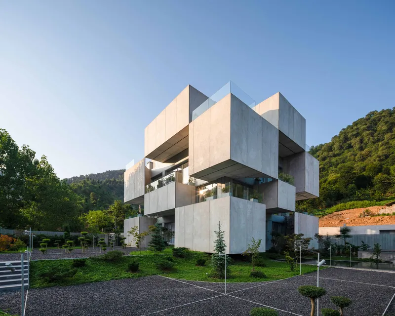 Stacked concrete volumes with cantilevered upper floors surrounded by lawn and young evergreens at dusk
