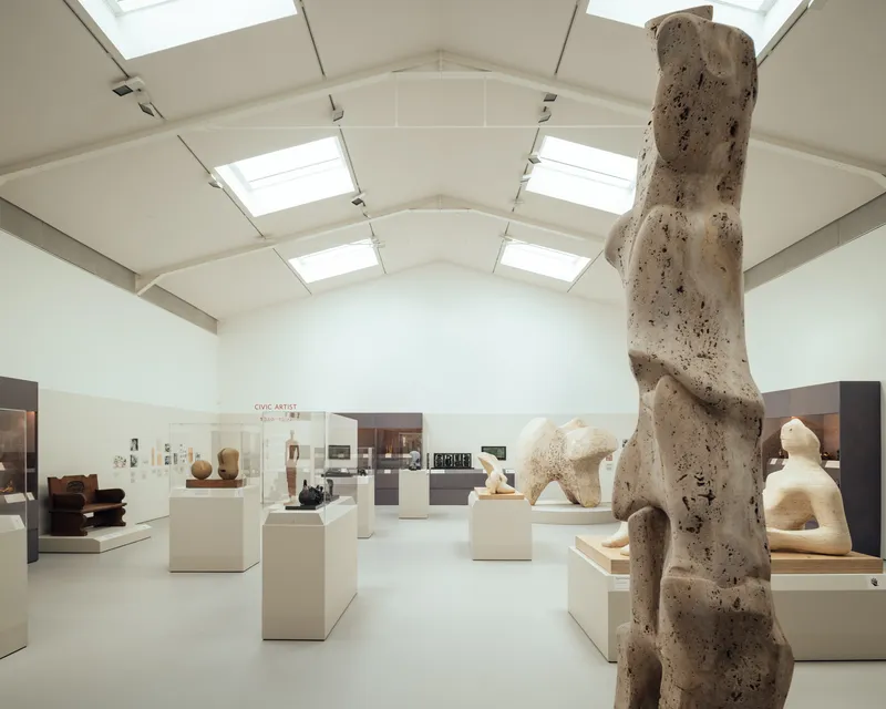 Gallery space with vaulted ceiling, skylights, and sculpture displays on white plinths