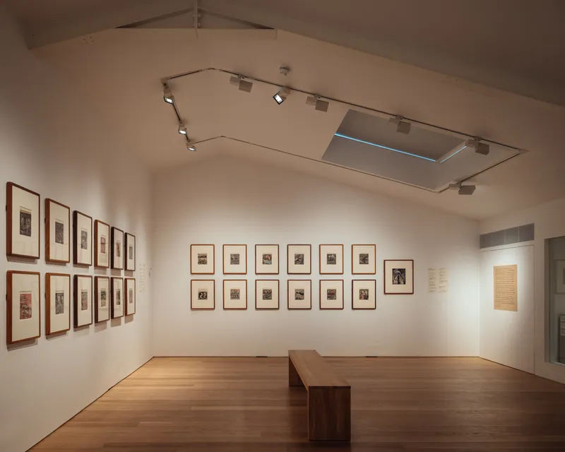 Exhibition room with framed artworks on white walls, timber floor, and recessed ceiling track lighting
