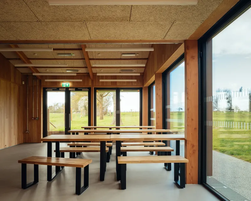 Communal dining area with timber benches and tables overlooking a grassy field through glazed doors