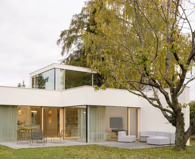 Two-story white volume with rooftop glazed pavilion and sliding glass doors opening to terrace beneath bare tree branches