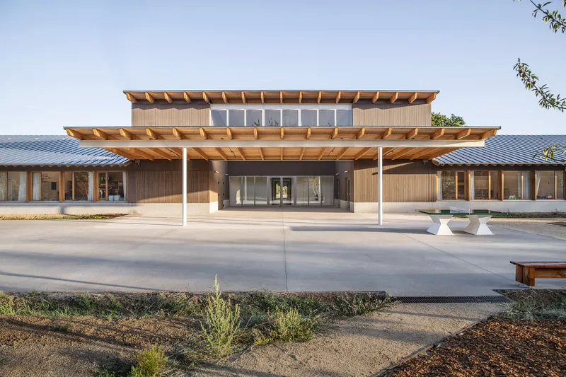 Main entrance with layered timber canopies and white columns sheltering a concrete plaza at dusk