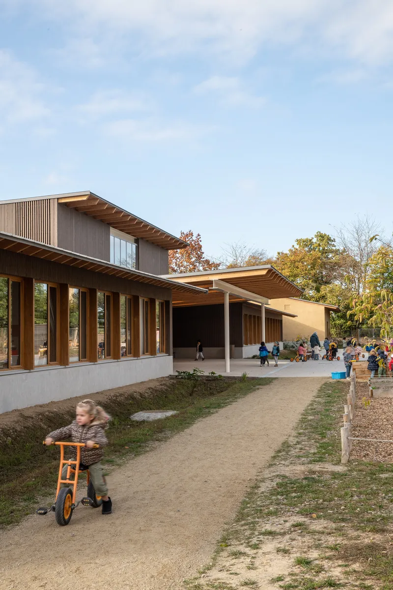 Exterior view with timber canopy over a concrete terrace where children play among autumn trees