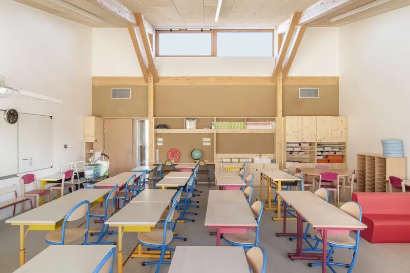 Classroom interior with exposed timber frame, clerestory windows, and rows of colorful desks under plywood ceiling
