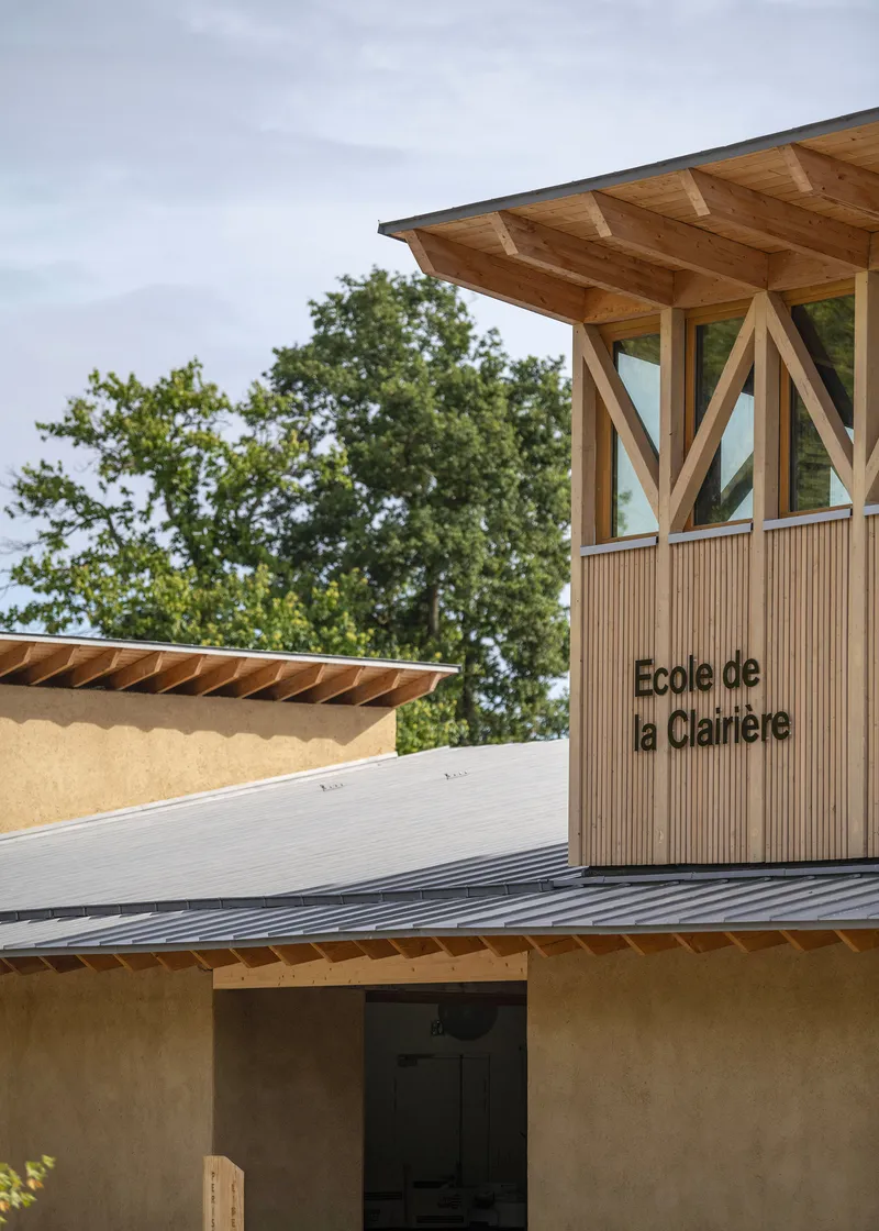 Entrance tower with timber bracing and slatted cladding rising above rammed earth volumes and metal roofing