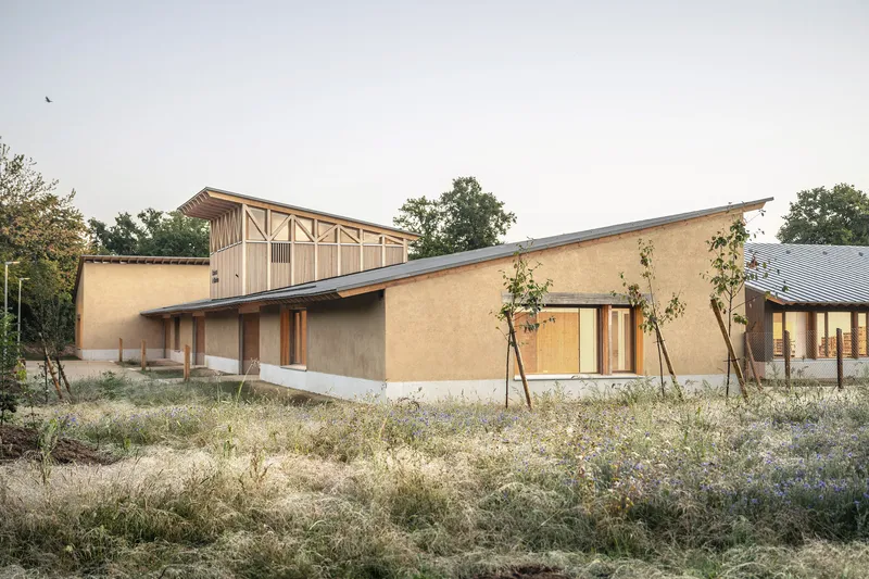 Earthen wall facade with timber columns and glazed openings beside raised timber structure at dusk