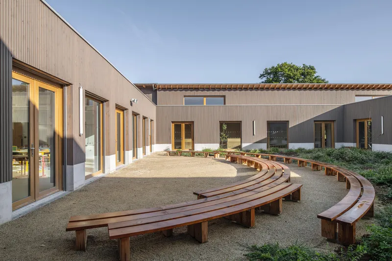 Central courtyard with curved timber benches on gravel facing timber-clad wings under clear sky