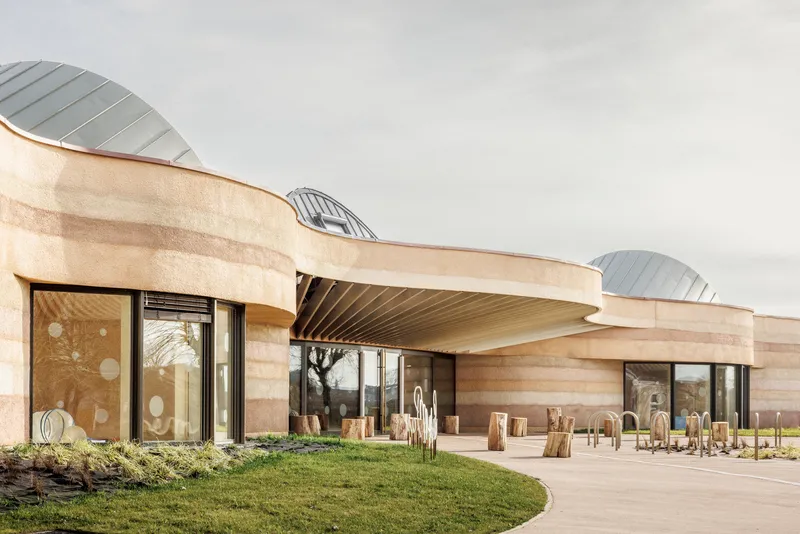 Entrance view of the rammed earth facade with curved overhangs and three undulating roof forms under an overcast sky