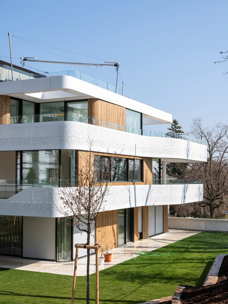 Garden facade with stacked balconies clad in perforated white panels and timber framing beneath winter trees