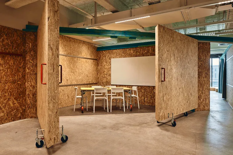 Movable oriented strand board partition panels on industrial castors defining a meeting room beneath exposed mechanical systems